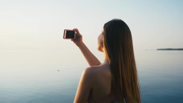 Pretty Young Woman Tomando fotos con su teléfono inteligente al atardecer en la playa cerca del mar de vacaciones. Moción lenta — Vídeos de Stock