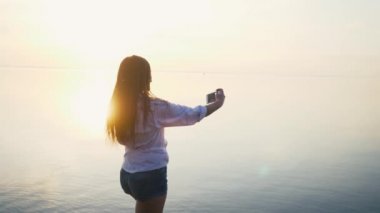 Tatile denize sıfır plaj üzerinde günbatımında onun Smartphone ile güzel genç kadın alarak resimler. Ağır çekim
