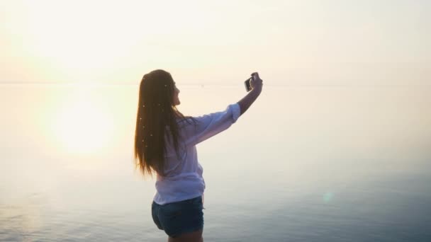 Jolie jeune femme prenant des photos avec son smartphone au coucher du soleil sur la plage près de la mer en vacances. Mouvement lent — Video