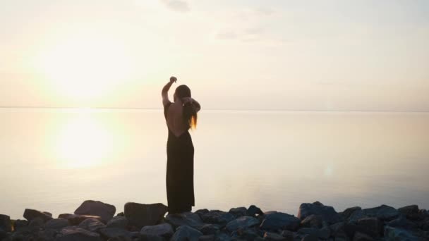 Mulher despreocupada em vestido preto longo dançando na praia ao pôr do sol — Vídeo de Stock