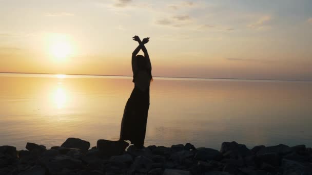 Беззаботная женщина в длинном черном платье танцует на пляже на закате — стоковое видео