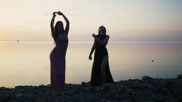 Mulheres despreocupadas em vestido preto longo dançando na praia ao pôr do sol — Vídeo de Stock