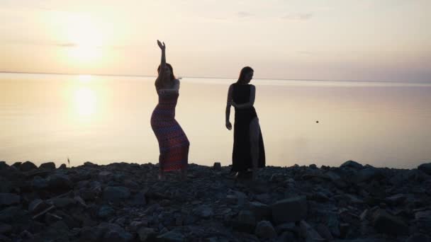 Mulheres despreocupadas em vestido preto longo dançando na praia ao pôr do sol — Vídeo de Stock