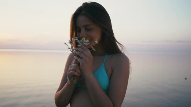 Молодая женщина стоит на берегу моря и нюхает цветок. Ношение бикини и шорт — стоковое видео