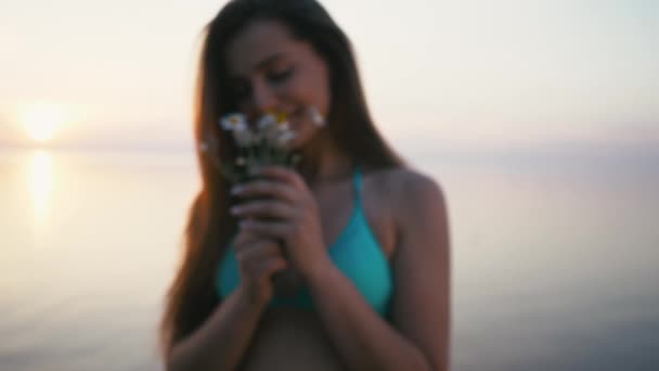 海岸に立っている花の臭いがする若い女性。ビキニとショート パンツを着てください。 — ストック動画