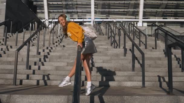 Ελκυστική χαριτωμένο νέοι Hipster Millenial γυναίκα φοιτητής έχοντας διασκέδαση ιππασία ένα κάγκελο και ακούγοντας μουσική στο αστικό φόντο. Φορώντας backpack, κίτρινη μπλούζα και ασημένια φούστα — Αρχείο Βίντεο
