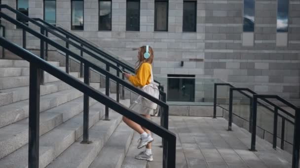 Retrato de joven linda y atractiva joven en las calles urbanas de la ciudad fondo escuchando música con auriculares. Mujer con blusa amarilla y falda plateada — Vídeos de Stock