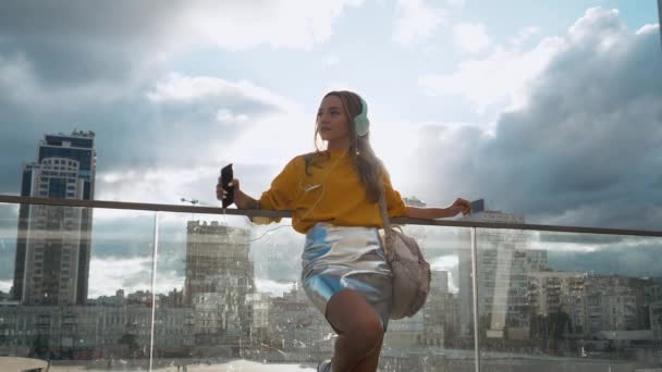 Porträtt av ung söt attraktiv ung flicka i urbana staden gator bakgrunden lyssnar på musik med hörlurar. Kvinna som bär gula blusen och silver kjol — Stockvideo