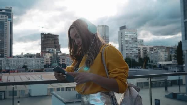 Portret van jonge leuke aantrekkelijke jonge meisje in stedelijke stad straten achtergrond muziek met een hoofdtelefoon beluisteren. Vrouw met gele blouse en zilveren rok — Stockvideo