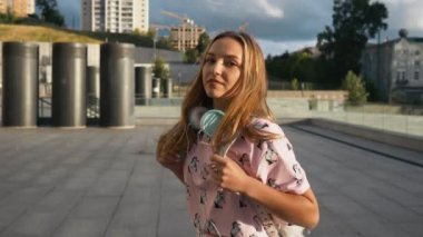 Hareket portre çekici sevimli gülümseyen beyaz elbise genç kadın kentsel ortamda sırt çantası ve kulaklık ile yavaş. Millenial Hipster yeni nesil Y