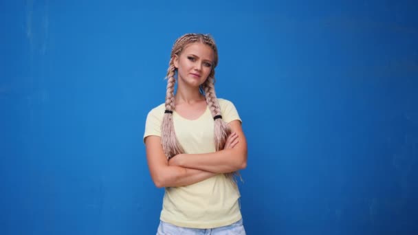 Mujer estudiante hipster rubia seria mirando a la cámara con los brazos cruzados sobre fondo azul — Vídeos de Stock