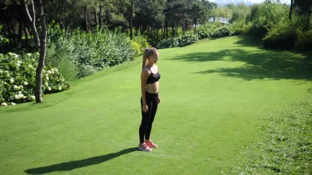 Mujer rubia joven con trenzas haciendo ejercicios de ejercicios de ejercicios de saltos en el prado en el verde parque de verano. Movimiento lento — Vídeos de Stock