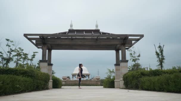 Joven mujer caucásica en ropa deportiva relajándose practicando yoga descalza en el parque de la ciudad. Fondo del amanecer. Movimiento lento — Vídeos de Stock