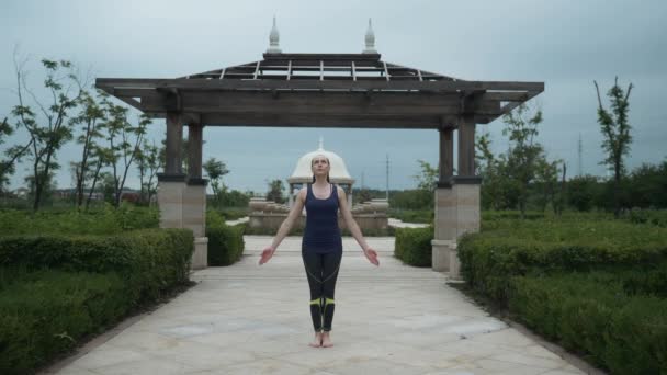 Joven mujer caucásica en ropa deportiva relajándose practicando yoga descalza en el parque de la ciudad. Fondo del amanecer. Movimiento lento — Vídeo de stock