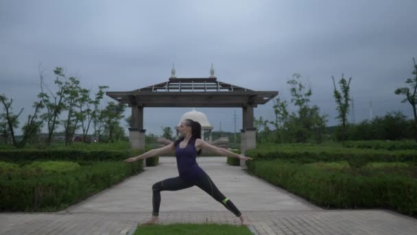 Joven mujer caucásica en ropa deportiva relajándose practicando yoga descalza en el parque de la ciudad. Fondo del amanecer. Movimiento lento — Vídeos de Stock