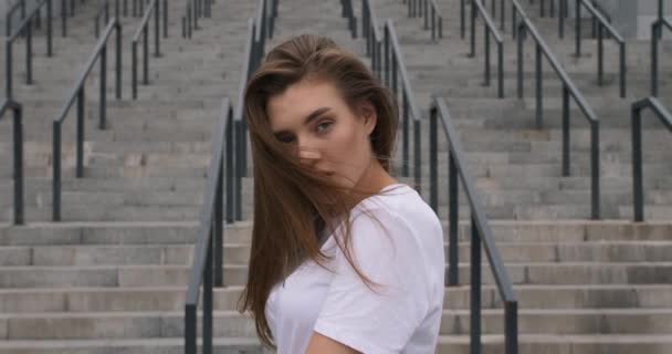 Retrato dramático de bela mulher caucasiana em camisa branca olhando sério câmera lenta suave luz natural no rosto ao ar livre na cidade. Escadas em segundo plano — Vídeo de Stock