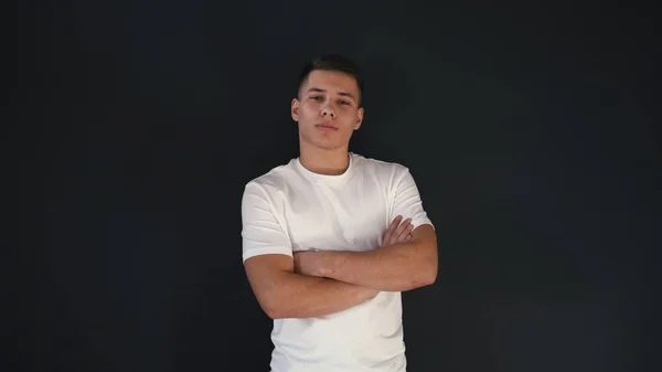Attractive and confident young man stands in white shirt and poses on camera with crossed hands. Guy looks very confident and good. Isolated on black background