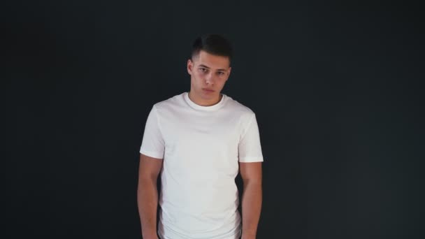 Tired young man stands in white shirts and looks on camera. He is serious. Then guy starts to smile and laugh. He moves his head and body a bit. Young man is emotional. Isolated on black background. — Stock Video