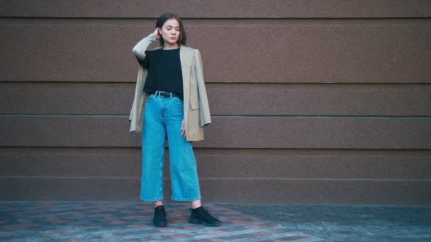 Портрет красивой модной смешанной расовой хипстерши в пиджаке и джинсах. Она позирует перед коричневой стеной. — стоковое видео