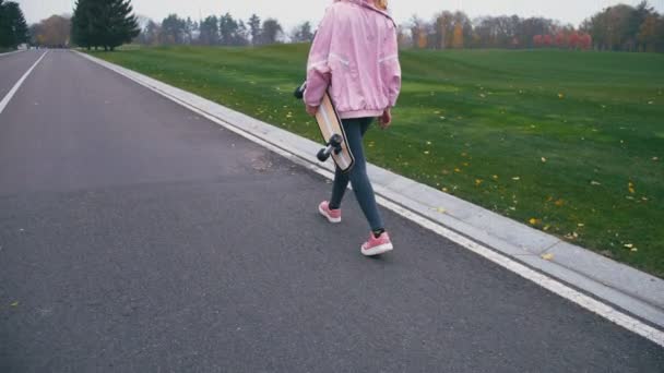 Young pretty beautiful blond hipster woman walking down the road with skateboard longboard in slow motion — Stock Video