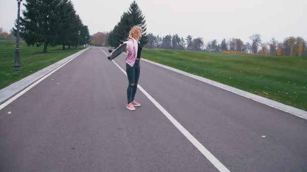 Молода красива блондинка хіпстерка, що йде по дорозі зі скейтбордом у повільному русі — стокове відео