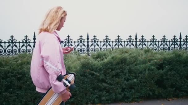 Vrij mooie blonde hipster vrouw met skateboard longboard met behulp van haar smartphone mobiele telefoon voor het doorbladeren, sms of chatten — Stockvideo