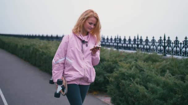 Довольно красивая блондинка хипстер женщина со скейтбордом на длинной доске, используя свой смартфон сотовый телефон для просмотра, смс или чата — стоковое видео