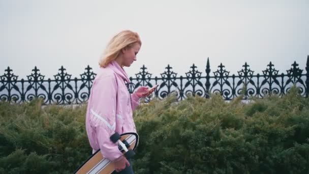 Mujer hipster rubia bastante hermosa con longboard monopatín utilizando su teléfono inteligente para navegar, SMS o chat — Vídeo de stock