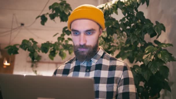 Hipster hombre en sombrero amarillo usando ordenador portátil y trabajando mientras está sentado en la cafetería o espacio de coworking — Vídeos de Stock