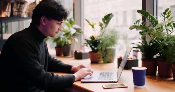 Hombre de raza mixta trabajando con el ordenador portátil y el teléfono inteligente en la cafetería — Vídeos de Stock