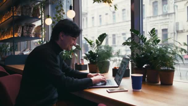 Hombre de raza mixta trabajando con el ordenador portátil y el teléfono inteligente en la cafetería — Vídeos de Stock