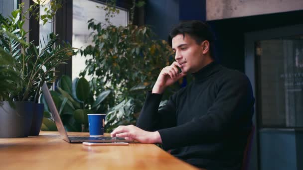 Hombre de raza mixta trabajando con el ordenador portátil y el teléfono inteligente en la cafetería — Vídeos de Stock