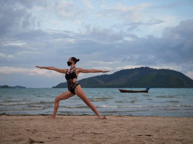 Kafkas kadın siyah mayo ve tıbbi Anti-smog maskesi giyen deniz kıyısı, Yoga uygulamak