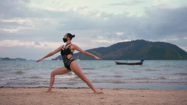Mujer caucásica practicando yoga en la orilla del mar con traje de baño negro y máscara médica anti-smog — Vídeos de Stock