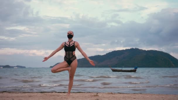 Mujer caucásica practicando yoga en la orilla del mar con traje de baño negro y máscara médica anti-smog — Vídeos de Stock