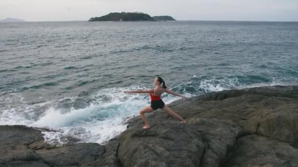 Spokojny kobieta praktykowania jogi fitness ćwiczenia na skalistej plaży przed burzliwy ocean — Wideo stockowe