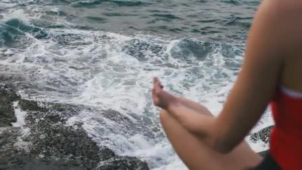 Спокійна жінка практикує фізичні вправи на скелястому пляжі перед штормовим океаном — стокове відео