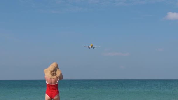 Туристична жінка в червоному купальнику і капелюсі вітає літак для подорожей. Привіт концепція відпустки — стокове відео