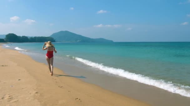 Lyckligt leende ung kvinna i solhatt och röda baddräkt promenader på stranden längs havet, havet semester och resa koncept — Stockvideo
