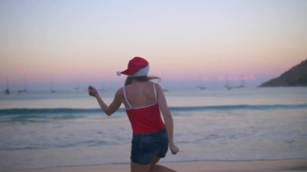 Unbekümmerte Frau tanzt und springt glücklich in Weihnachtsmütze und rotem Badeanzug bei Sonnenuntergang am Strand des tropischen Paradieses — Stockvideo