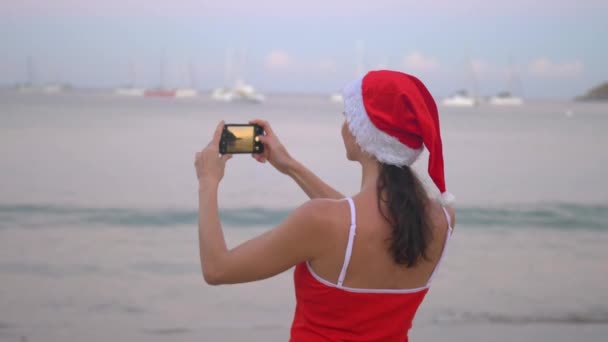 日出时, 戴着圣诞老人帽子和红色泳衣的妇女在海边用智能手机移动相机拍照 — 图库视频影像
