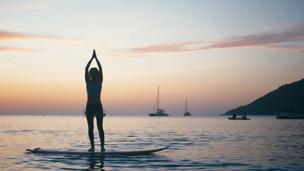 Young woman doing yoga on sup board with paddle at sunset