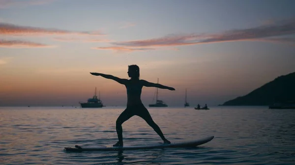 Young woman doing yoga on sup board with paddle at sunset