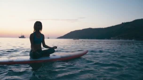 日落时, 年轻女子在冲浪板上用划桨做瑜伽 — 图库视频影像