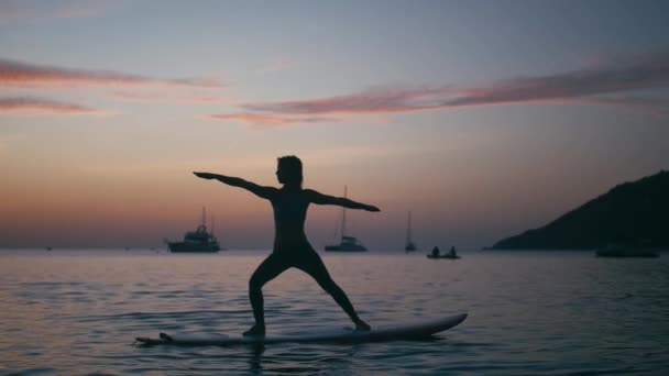 Jeune femme faisant du yoga sur une planche de sup avec pagaie au coucher du soleil — Video