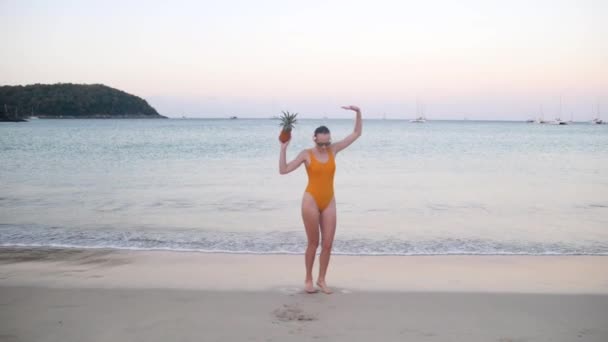 Mujer en traje de baño amarillo divirtiéndose bailando con piña en la playa al atardecer — Vídeos de Stock