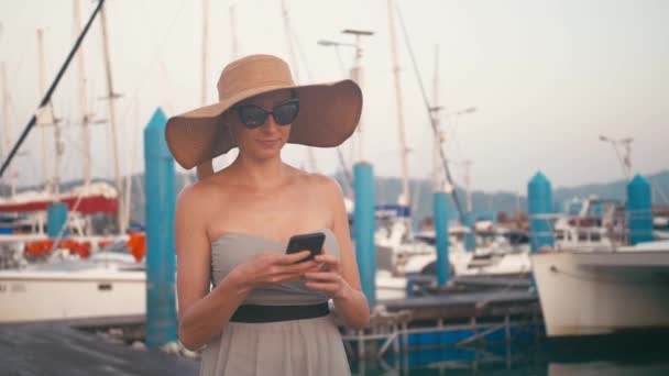 Dame chic en robe en utilisant son smartphone marche dans la jetée de la marina avec des yachts à l'arrière-plan — Video