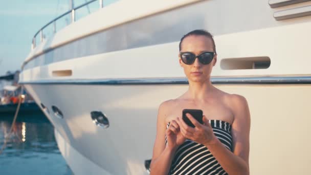 Dame chic en robe en utilisant son smartphone dans la jetée de la marina avec des yachts à l'arrière-plan — Video