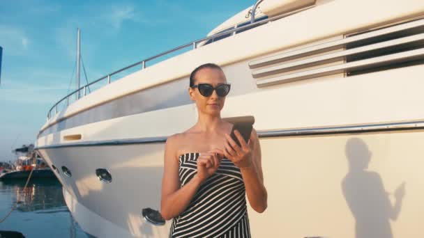 Dame chic en robe en utilisant son smartphone dans la jetée de la marina avec des yachts à l'arrière-plan — Video