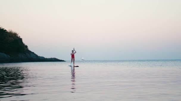 Mujer en traje de baño rojo en SUP de pie tabla de paddle en una bahía al atardecer — Vídeos de Stock